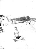 Badklädda människor på en strand, i förgrunden en man iförd badbyxor och solglasögon. I bakgrunden syns barrskog.