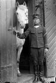 En man i soldatuniform står i en stalldörr där en vit häst tittar ut.