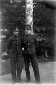 Två män i soldatuniform står på en väg framför ett träd i skogen.