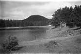 I skogskanten vid en sjö ligger en liten stuga och en båt ligger uppdragen på stranden. I bakgrunden höjer sig en skogsklädd kulle.