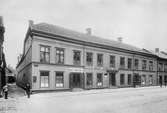 År 1890. Vid Östra Storgatan i Jönköping ligger Gustaf Holms juridiska byrå. Bredvid finns Amelie Samuelsson & Co och vid andra sidan G.W.Rehdahl.
I fönstren på andra våningen står två mustaschprydda män och tittar ut.
I gränden sitter en skylt med texten: Pensionat, in på gården, en trappa upp.