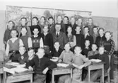 Pojkar sittandes i bänkar och stående bakom i klassrummet på Kvarnbyskolan (f.d. Centralskolan). Magister Sigfrid Eyton med elever.