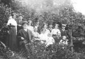 Släktfoto där män, kvinnor och barn sitter och står utomhus i en slänt vid en träddunge, cirka 1900-talet. Okänd plats.