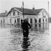 Uniformsklädd man (Polisman) under tjänstgöring vid översvämning på Göteborgsvägen i Mölndalsbro. Göteborgs- och Bohusläns sparbank syns i bakgrunden. Omkring 1920