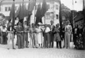 Flertalet män och kvinnor, medlemmar från SSU-föreningen i Mölndal. De står med flaggor, troligtvis politiskt möte, på torget i Halden. 1930-tal.