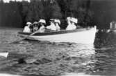 Sju damer med vackra hattar i en roddbåt på Rådasjön, 1910-07-17. I båten sitter bland andra Ester Alberts, mor till givaren Gunnel Dahlbeck. 
I bakgrunden ses  