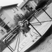 Ett litet barn ståendes i karusell i en lekpark, 1965. Brunnsgatan 4 och 6 syns i bakgrunden.