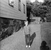 En pojke står på gruset utanför huset. I bakgrunden intill väggen syns en trehjuling. Brunnsgatan 9, 1965