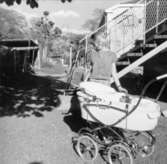 En flicka, Connie Olsén, står vid en barnvagn utanför husen på Brunnsgatan. 1960-tal.