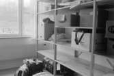 Dokumentation av Sagåsens flyktingförläggning 1992. Ett rum med tre ihopmonterade lagerhyllor som innehåller kartonger, resväska och kassar. I bakgrunden finns fönster.