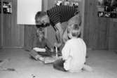 Två barn sitter på golvet och håller i ett träföremål. En vuxen hjälper dem. Utställningsvernissage av och om Katrinebergs daghem på Mölndals museum 1993-09-10.