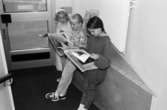 Tre flickor står lutade mot en entrévägg och läser tidningar. Utställningsvernissage av och om Katrinebergs daghem på Mölndals museum 1993-09-10.