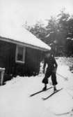 Okänd man åker skidor utför en sluttning invid ett hus i Fjärås Bräcka, 1940-tal.