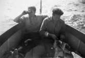 Helmer Garthmans arbetskompisar är ute med båt och fiskar. En man tittar i kikare utöver vattnet och den andre rensar fisk på ett tidningspapper. Näset, 1950-tal. Skänkt av sonen Alf Garthman som arbetade under många år på Mölndals stadsmuseum.