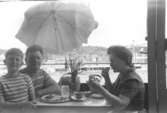 Familjen Garthman sitter på balkongen och fikar. Från vänster Jerry, Helmer och Astrid på Lerbrännargatan 1F, 1963. I bakgrunden skymtar lyftkranar och nybygget av Idrottshuset (senare Aktiviteten). Bygget pågick 1962-1965.