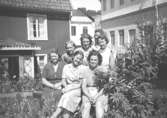 Sju kvinnliga arbetskamrater (okända för givaren) står/sitter i gräset framför ett hus och en fabrik. De är från 