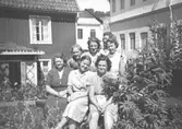 Sju kvinnliga arbetskamrater (okända för givaren) står/sitter i gräset framför ett hus och en fabrik. De är från 