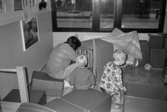 En förskollärare vid Katrinebergs daghem och två små barn bygger en koja med hjälp av filtar över en barnsäng. I sängen sitter några barn. Omkring dem finns lekbackar och fyrkantiga sitt-kuddar.