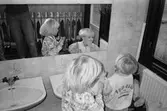 Två små barn står framför en spegel och tvättar sina händer och mun i handfatet. I bilden speglas fröken som står strax bakom. Väggen bakom handfatet är kaklat med bl.a blom-dekor. Katrinebergs daghem.