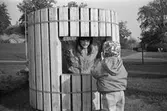 Dagisbarn som leker utomhus vid Katrinebergs daghem. En flicka står inne i en rund, hög (ca 1 meter), smal trätunna som är omgärdad av spännband i stål. Det liknar en slags enkel kiosk, gjord av långsmala högstående ribbor, med ett fyrkantigt stort hål i mitten, så att man kan titta ut. Utanför står en liten flicka, klädd i byxor, jacka och blommig mössa. Hon tittar på den lite större flickan innanför.