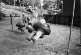 Tre barn gungar i en gungställning som står i en stor sandlåda. I bakgrunden står en fröken med ett annat barn. Längre bort ser man stora däck man kan klättra i. Katrinebergs daghem, 1992.