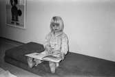 En liten flicka, barfota och iklädd mjukiskläder, sitter på ett par madrasser och tittar mot fotografen. Hon bläddrar i en bok som ligger på hennes ben. I bakgrunden till vänster hänger en inglasad tavla med ett troll på. Katrinebergs daghem, 1992.