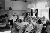 En fröken och fem barn sitter i köket runt ett furubord och fikar. En förskollärare står vid diskbänken och pysslar med något. De sitter på både pinnstolar och bruna eller träfärgade tripp trap.
