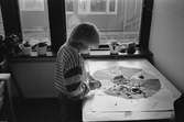 En pojke står vid ett bord som är uppställt brevid ett fönster. På bordet ligger en teckning som är målat i olika kulörer som en delad apelsin. Barnet målar med en pensel i en av 