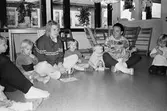 Två förskollärare, varav en är Helén Johansson, och flertalet småbarn sitter tillsammans i en cirkel på golvet. En lärare har ett barn i sitt knä. Kullerbyttan, Katrinebergs daghem, 1992.