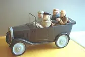 Fyra personer åker i en bil. Två personer sitter fram och två bak. Bilen, modell T-Ford, är mörkgrå och saknar tak. Däcken är ljusgrå. Man ser ekipaget snett från sidan. Hör ihop med bild nr 1994_0959. Harry Bergmans 