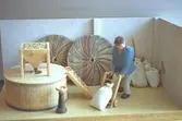En manlig träfigur, iklädd keps, går bredvid en mjölkvarn. Framför sig skjuter mannen en transportkärra, lastad med en fylld säck. Kvarnen består av en rund tunna, ovanpå ligger ett kvarnhjul samt en behållare för korn. Från kvarnhjulet finns ett utlopp för det som är färdigmalt. I bakgrunden ser man fyllda mjölsäckar, borstar och två kvarnhjul. Harry Bergmans 