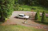En vit, bil (Saab) står parkerad vid några träd, Gunnebo slott.