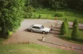 En vit, bil (Saab) står parkerad vid några träd, Gunnebo slott.