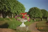 En vit blomsterurna fylld med färgglada blommor i. Urnan står på en piedestal av sten. Vid sidan om löper en grusstig och fint klippta träd.