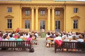 Människor som sitter och tittar på en teaterföreställning på framsidan av Gunnebo slott. En skådespelare, iklädd en svart hatt och bar överkropp, håller händerna ovanför en grön soptunna. Till vänster står en anann skådespelare i orange overall.