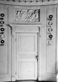 Utsmyckad enkeldörr med relief ovan. På båda sidor om dörren hänger koppar och fat av porslin. Gunnebo slott, 1930-tal.