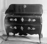 Byrå av mörkt träslag med mässingsbeslag, två lådor och framåtlutande överdel. Gunnebo slott 1930-tal.