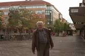 En äldre man står på Brogatan. I bakgrunden till vänster ses Alberts torg och Havskatten 17. Mölndalsbro i dag - ett skolpedagogiskt dokumentationsprojekt på Mölndals museum under oktober 1996. 1996_1301-1319 är gjorda av högstadieelever från Åbyskolan 8A, grupp 4. Se även 1996_0913-0940, gruppbilder på klasserna 1996_1382-1405 och bilder från den färdiga utställningen 1996_1358-1381.