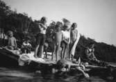 En kvinna och några barn sitter eller står på en flotte som ligger i Tulebosjön, 1930-tal. Framför flotten står två kvinnor som håller var sin kaffekopp i handen.