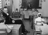 Biskop Bo Giertz besöker Brattåsskolan och sitter bland eleverna i ett klassrum 14 feb 1955. Vid katedern sitter Lärarinnan Greta Andersson (född Alm).