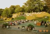 Publik som lyssnar till en pianokonsert sitter både på kullen samt uppställda bänkar. Gunnebo slotts framsida, augusti 1990.