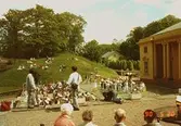 Till vänster ses en stående man som filmar en sångkör och dess publik, vilka sitter på gräsmattan eller står framför Gunnebo slott, maj 1990.