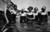 Sex badklädda kvinnor står i vattnet, troligtvis i Tulebosjön cirka 1930. Till vänster ses en träbyggnad.