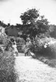 Sommar i Lindome i början av 1940-talet. En flicka och en kvinna står vid en grind i en trädgård.