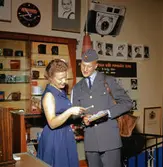 Brevbärare iklädd postens uniform från 1962 års
beklädnadsreglemente, överlämnar post till kvinnlig expedit i en
fotobutik. Brevbäraren på bilden är med stor sannolikhet Owe Jonsson.
Owe Jonsson var brevbärare i Växjö och vann EM-guld i Belgrad den 16
september 1962 i löpning 200 meter, bara 13 dagar senare omkom han i
en bilolycka.