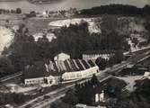 Inramat flygfoto över Forsåkers fabriker vid Grevedämmet i Mölndal. I bakgrunden ses Stensjön och Stensjöhill. Fabriksbyggnaden kallades tidigare för Bettyholm och Carlsfors fabriker. Relaterat motiv: 2012_0171.