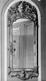Vägghängd, avlång spegel, rundad upptill och med dekor. Spegeln är insatt i ett valv och glaset är i två delar, därav diffus spegelbild. Gunnebo slott 1930-tal.