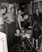 Översvämning på varuhuset Nordiska Kompaniet i Stockholm 1947. Fyra män, troligen brandmän, står runt en teknisk apparat med slangar och mätare. Mannen som sköter apparaten är klädd i uniformsjacka och mössa, hans byxor är uppkavlade. De övriga männen har bar överkropp, en av dem är tatuerad. I bakgrunden står en något äldre man i kavaj, skjorta och slips. Det är vatten på golvet.