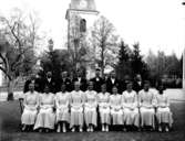 Konfirmander, 10 flickor, 7 pojkar och kyrkoherde Ivar Jäderlund.
Rinkaby kyrka i bakgrunden.