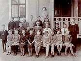 Rynninge folkskola, 23 skolbarn med lärare fru Dahlin.
Skolbyggnad i bakgrunden.